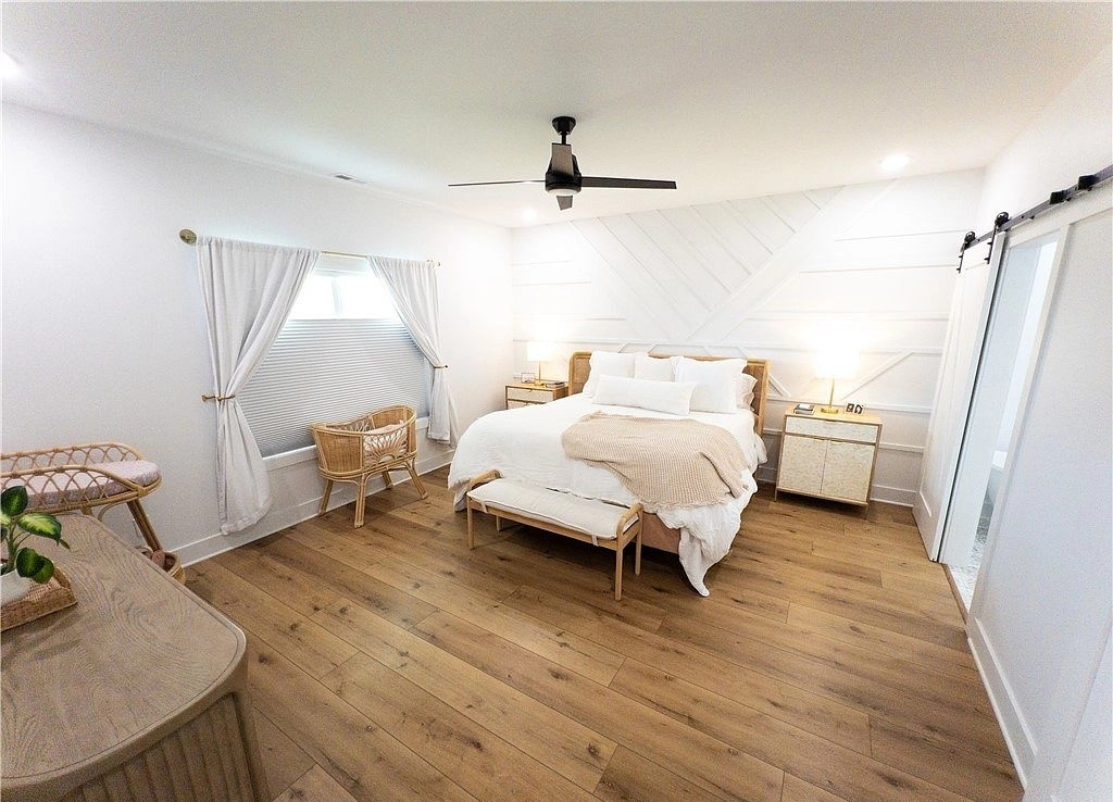 Furnished bedroom with wood flooring, white curtains, and a sliding bathroom door.