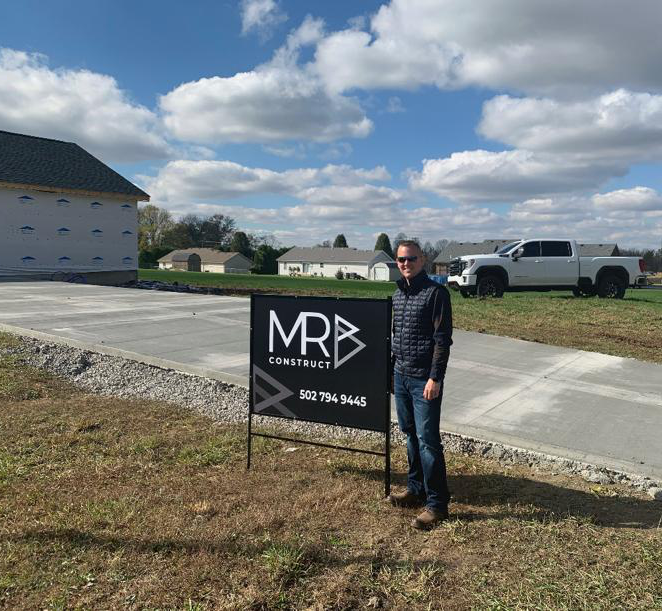 Michael Branham standing beside an MRB Construct sign at a home-building site